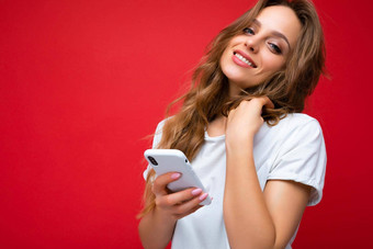 漂亮的可爱的年轻的金发女郎女人穿白色t恤站孤立的红色的背景冲浪互联网电话相机