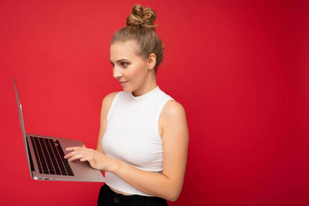 一边配置文件照片拍摄美丽的金发碧眼的年轻的女人聚集头发穿白色t恤电脑移动PC打字键盘上网本监控屏幕孤立的红色的墙背景