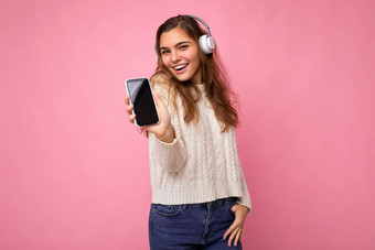 美丽的快乐微笑年轻的女人穿时尚的休闲装孤立的背景墙持有显示移动电话空显示模型穿白色<strong>蓝牙耳机</strong>听音乐有趣的相机