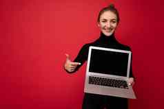 照片美丽的微笑快乐年轻的浅黑肤色的女人女人持有电脑移动PC空监控屏幕穿黑色的长袖相机的印刷上网本孤立的红色的墙背景
