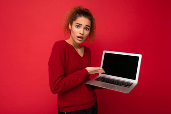 肖像美丽的惊讶惊讶惊讶黑暗金发碧眼的女人持有移动PC电脑相机显示上网本键盘穿红色的毛衣孤立的红色的墙背景