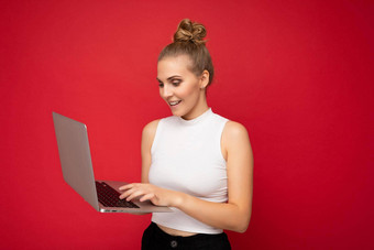 一边配置文件照片拍摄美丽的微笑快乐金发碧眼的年轻的女人聚集头发穿白色t恤电脑移动PC打字键盘上网本监控屏幕孤立的红色的墙背景