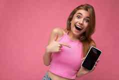 照片美丽的快乐快乐的年轻的金发女郎女人穿粉红色的前平衡孤立的粉红色的背景空空间持有手显示移动电话空显示模型相机指出手指gadjet