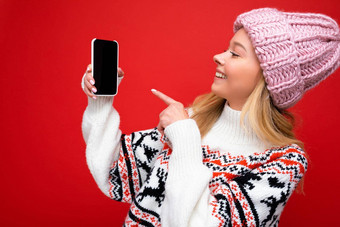 照片有吸引力的微笑年轻的金发女郎女人穿温暖的针织他冬天温暖的毛衣站孤立的红色的背景显示智能手机空屏幕模拟设备显示指出手指gadjet