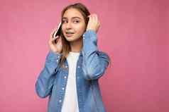 照片美丽的深思熟虑的年轻的金发女郎女人穿休闲蓝色的珍衬衫孤立的粉红色的背景持有手会说话的移动电话相机思考