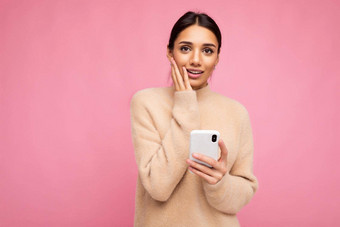 有吸引力的可爱的伤心年轻的褐色女人穿米色温暖的毛衣站孤立的粉红色的背景冲浪互联网电话相机有关