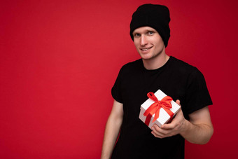 拍摄英俊的快乐年轻的男人。孤立的红色的背景墙穿黑色的他黑色的t恤持有白色礼物盒子红色的丝带相机