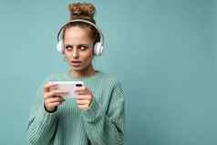 特写镜头照片美丽的伤心年轻的女人穿蓝色的毛衣孤立的蓝色的背景穿白色蓝牙无线耳机听音乐移动电话沟通在线互联网一边理解