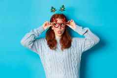 圣诞节聚会，派对庆祝活动概念愚蠢的红色头发的人女孩享受一年穿有趣的眼镜头巾显示舌头盯着左标志蓝色的背景