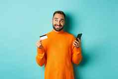 在线购物英俊的男人。思考持有智能手机信贷卡支付互联网商店站光蓝色的背景