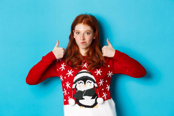 冬天假期圣诞节夏娃概念深刻的印象红色头发的人女孩圣诞节毛衣点头批准显示拇指赞美好产品站蓝色的背景