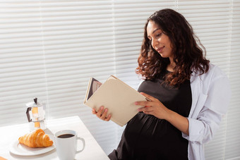 积极的怀孕了女人读取书早....早餐咖啡羊角面包概念愉快的早....期望会议婴儿