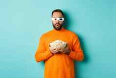 英俊的男人。橙色毛衣眼镜看电影激动持有爆米花惊讶站蓝色的背景