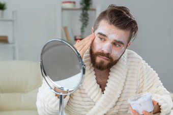 有胡子的的家伙应用面部奶油前面镜子皮肤护理水疗中心男人。概念