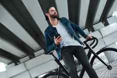 在户外休闲年轻的时尚的男人。坐着自行车城市街持有智能手机微笑快乐的