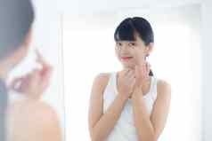 美丽的年轻的亚洲女人镜子应用保湿霜化妆品奶油乳液浴室首页护肤品卫生治疗身体护理皮肤抗衰老面部
