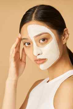 工作室拍摄年轻的女人面部面具应用一半脸相机接收水疗中心治疗孤立的米色背景