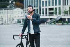 在户外休闲年轻的时尚的男人。走城市街自行车持有杯喝热咖啡深思熟虑的