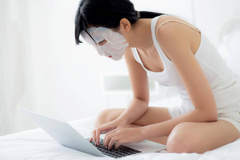 美丽的年轻的亚洲女人表面部面具坐着工作移动PC电脑床上卧室美女孩应用脸面具护肤品皱纹首页皮肤护理健康健康