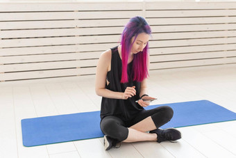 漂亮的年轻的混血女孩紫色的头发计数燃烧卡路里应用程序智能手机坐着体育席健身房概念瑜伽健身