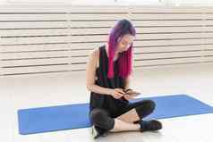 漂亮的年轻的混血女孩紫色的头发计数燃烧卡路里应用程序智能手机坐着体育席健身房概念瑜伽健身