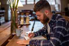 年轻的有胡子的商人坐在咖啡馆首页表格写笔记本谎言平板电脑电脑男人。工作研究
