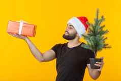 英俊的年轻的男人。持有红色的礼物盒子圣诞节树手摆姿势一年的帽黄色的背景快乐圣诞节快乐一年概念