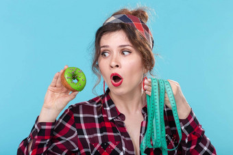 特写镜头震惊了惊讶年轻的美丽的女人测量磁带持有绿色甜甜圈手摆姿势蓝色的背景概念饮食拒绝有害的高热量的食物