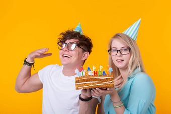 有趣的年轻的夫妇纸帽蛋糕使愚蠢的脸快乐生日站黄色的背景概念祝贺你愚弄