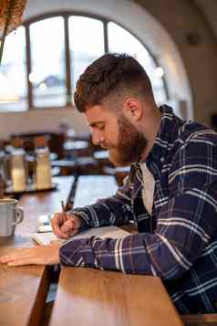 年轻的有胡子的商人坐在咖啡馆首页表格写笔记本谎言平板电脑电脑男人。工作研究