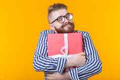 快乐的年轻的赶时髦的人的家伙胡子眼镜拥抱红色的礼物盒子黄色的背景概念快乐礼物