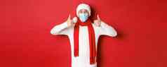 概念一年冠状病毒假期快乐的男人。庆祝一年社会距离穿医疗面具圣诞老人他围巾显示竖起大拇指批准