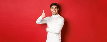 概念冬天假期圣诞节生活方式肖像快乐的有吸引力的男人。白色毛衣显示竖起大拇指批准好的想法站红色的背景