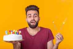 积极的年轻的男人。持有快乐生日蛋糕燃烧孟加拉灯摆姿势黄色的背景