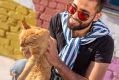 宠物老板友谊概念英俊的阿拉伯男人。持有拥抱可爱的姜猫猫好奇的表达式特写镜头