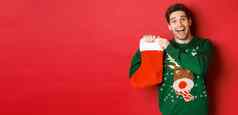 图像有吸引力的快乐的男人。毛衣持有圣诞节长袜礼物庆祝冬天假期站红色的背景