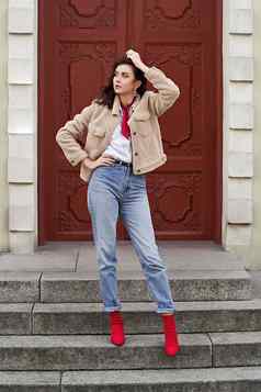 年轻的美丽的女人红色的高非常鞋子蓝色的牛仔布牛仔裤裤子泰迪夹克外套摆姿势红色的通过背景欧洲城市