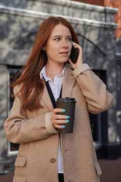 高加索人女人时尚的米色外套调用智能手机喝咖啡