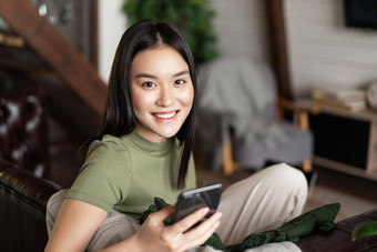微笑亚洲女人坐着沙发上首页舒适的衣服移动电话应用程序快乐相机