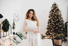 迷人的怀孕了女人提出了相机白色衣服圣诞节树lor灯
