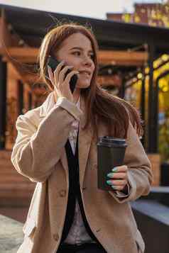 高加索人女人时尚的米色外套调用智能手机喝咖啡