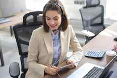 快乐的女经理坐着办公室桌子上执行企业任务无线连接数字小工具