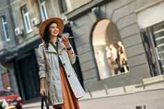 年轻的美丽的时尚的女人穿秋天外套他会说话的智能手机微笑走城市街道