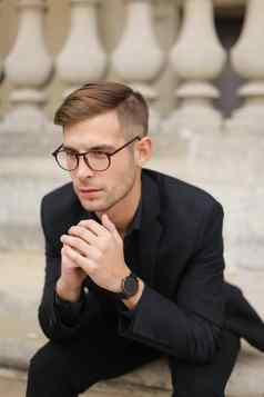 年轻的时尚的男人。眼镜坐着人行道上倾斜混凝土栏杆上