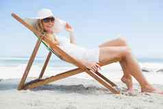 华丽的金发女郎坐着海滩穿遮阳帽太阳镜