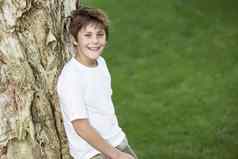 肖像可爱的十几岁的男孩倾斜树树干草坪上