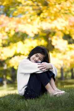 女孩拥抱膝盖坐着草坪上明亮的秋天叶子背景