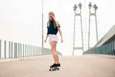年轻的美丽的金发女郎女孩骑明亮的滑板桥