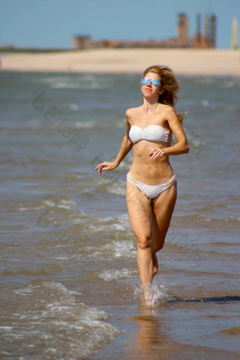 漂亮的晒黑金发女郎女孩白色比基尼太阳镜运行水桑迪海滩波罗的海海海岸