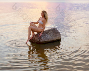 美丽的金发女郎白色比基尼坐在石头站水日落